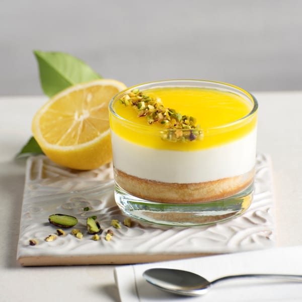 Lemon cheesecake in a glass, topped with pistachios, next to a lemon half and spoon.