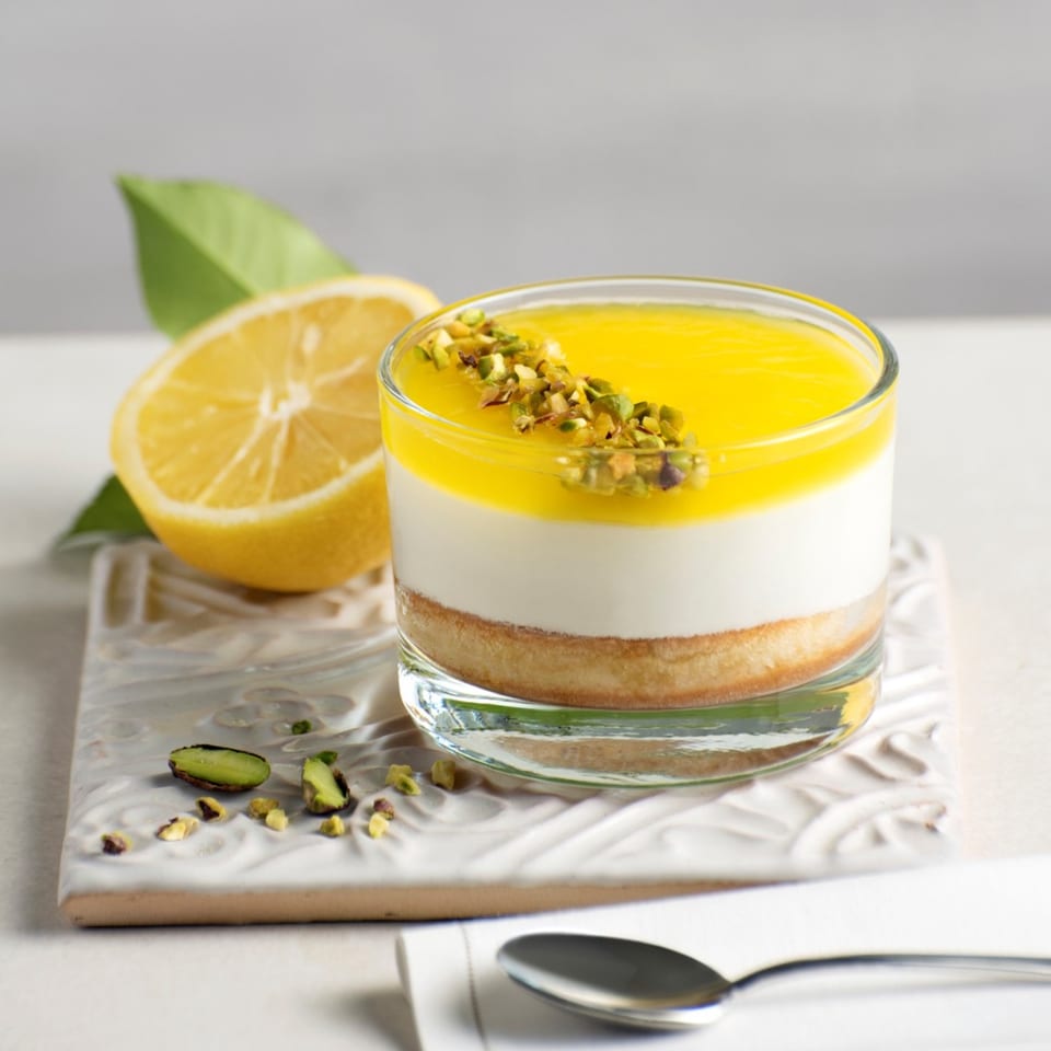 Lemon cheesecake in a glass, topped with pistachios, next to a lemon half and spoon.