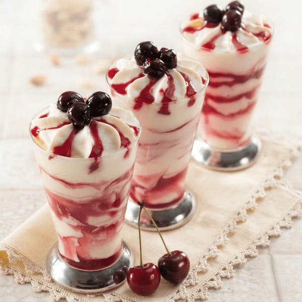 Three cherry parfaits with whipped cream and cherry syrup, garnished with fresh cherries on a lace napkin.