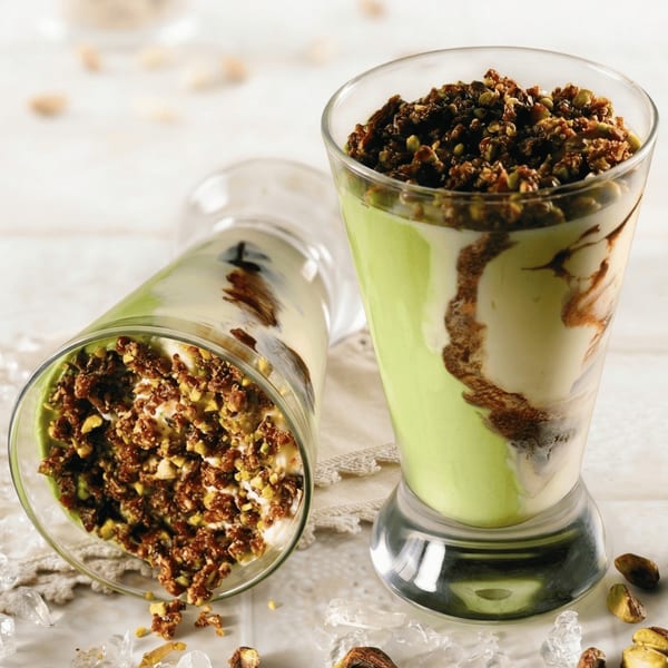 Two glasses of pistachio and vanilla dessert with chocolate swirls and nut topping, one upright, one tilted.