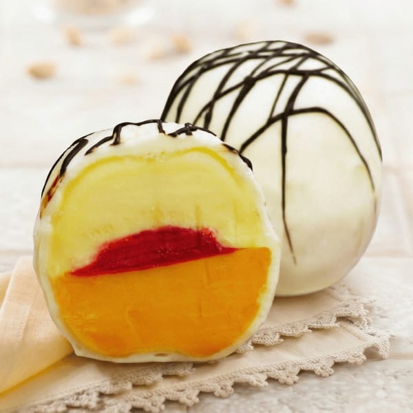 A sliced dessert ball reveals layers of yellow, red, and orange, with another whole dessert in the background.
